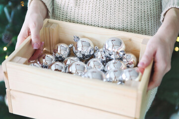 Woman holds a box with a lot of Christmas silver decorations near to the Christmas tree