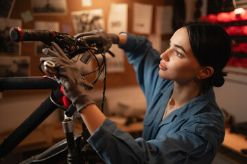 Close-up portrait of concentrated cycling mechanic woman checking and repairing bicycle handlebar with tools while working in bike repair workshop with authentic interior. - obrazy, fototapety, plakaty