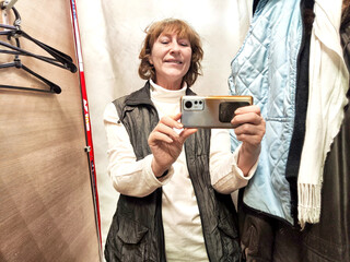 A middle-aged blonde woman in a cramped fitting room of a store with a bunch of clothes and hangers...