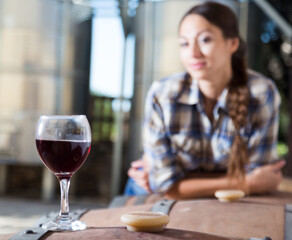 Girl looking at glass of red wine standing on wooden barrel in winery