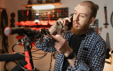 Man mechanic working in bicycle repair shop or garage, call on mobile phone, technician repairing bike using special tool.