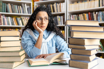 A thoughtful and focused woman sits among a pile of books in a library, reading and contemplating...