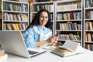A cheerful woman wearing glasses interacts with her smartphone while seated in a library surrounded by books. She exudes a sense of happiness and content in an academic setting.