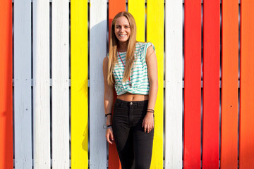 Stylish beautiful young woman poses in front of a vibrant colorful wooden wall on street, outdoors...