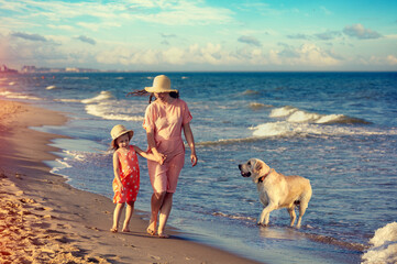 Young happy mother with little daughter holding hands and running barefoot on the beach. The dog runs nearby. Mother's Day concept.