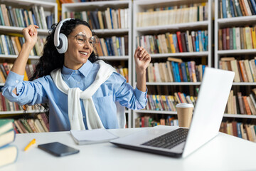 Excited young woman with headphones dancing joyfully at a library desk, surrounded by books. She exudes positivity and a love for music while working.