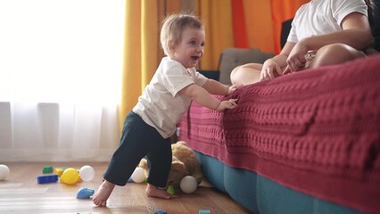 baby tries to get up, takes his a first steps, leans on the sofa. happy family kid dream concept. baby takes his first steps gets up lifestyle from his knees. baby takes first steps indoors