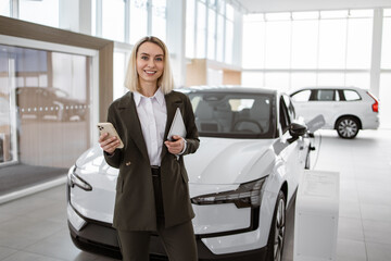 Portrait of female salesperson in car showroom. Attractive young woman standing in front of car...