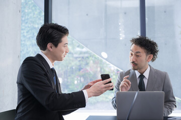 A businessman in a meeting room showing his phone and his boss instructing him on compliance so he doesn't catch on fire.