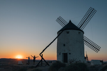 A windmill is standing in the distance with the sun setting behind it, two unrecognizable persons...