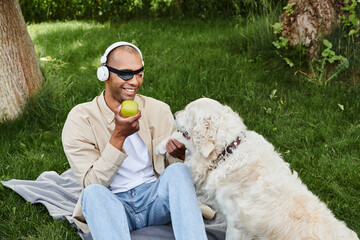 A disabled African American man with myasthenia gravis syndrome sits on a blanket with a Labrador dog, enjoying an apple.