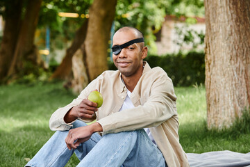 Naklejka na ściany i meble A disabled African American man peacefully sits on a blanket, cradling an apple in his hands
