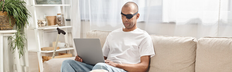 An African American man with Myasthenia Gravis syndrome is sitting on a couch, using a laptop, with...