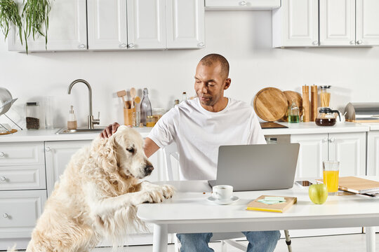 A disabled African American man sits at a table with a laptop, accompanied by a loyal Labrador dog.