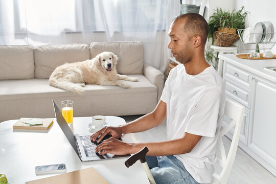 A disabled African American man sit at a table using a laptop computer, accompanied by a Labrador dog.