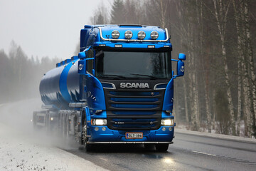 Obraz premium Blue Scania R580 tanker truck drives along wet road on a foggy day in winter to pick up a load.