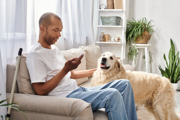 An African American man with myasthenia gravis sits on a couch, near his Labrador dog and using smartphone