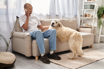 An African American man sits on a couch, talking on a cell phone next to his loyal Labrador dog at home.