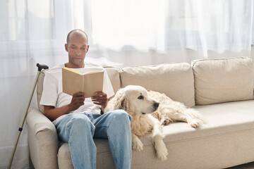 An African American man with myasthenia gravis is sitting on a couch with his Labrador dog, engrossed in reading a book.