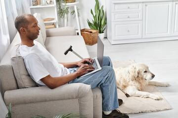 An African American man with myasthenia gravis, sitting on a couch, using a laptop beside his...