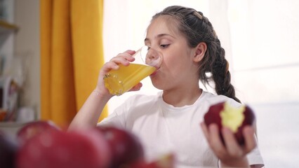 the child eats an apple and drinks juice. healthy eating fruit concept. child brunette girl eating an apple on the table in lifestyle the kitchen drinking apple juice indoors. kid drinks juice