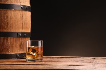 Whiskey with ice cubes in glass and barrel on wooden table against black background, space for text