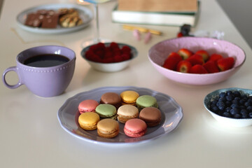 Plate of pastel macarons, cookies and chocolate, cup of tea of coffee, glass of bubble water, various berries, books and accessories on the table. Selective focus, pastel colors.
