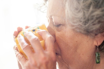 Seventy-year-old woman holding a cup of coffee with her hands while sipping it.