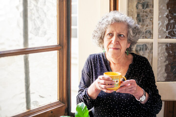 Smiling senior woman holds cup of coffee or tea to a window in her home.