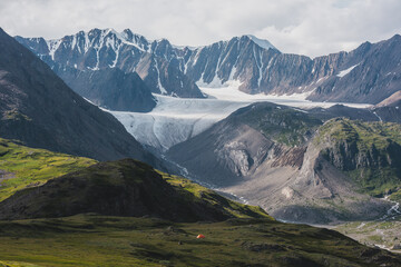 Dramatic landscape with orange tent in alpine valley with view to big glacier tongue and large sharp snow-capped mountain range under gray cloudy sky. Green hills and rocks against ice and sheer crags - 786185185