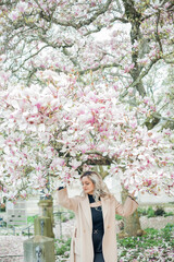 Beautiful young blonde with bouquet of flowers. Portrait of young woman near blooming magnolia in European city. Warm bright spring.