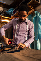 Male fashion tailor working on the suit design in his workshop cutting fabric with scissors.