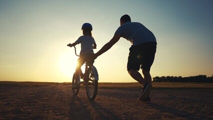 family play in the park. father teaching daughter to ride a bike. happy family kid dream concept. daughter learn to ride a bike silhouette. father supporting sunset child riding bike summer in park - obrazy, fototapety, plakaty