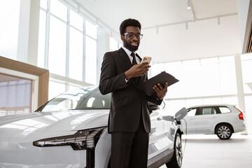 Smiling salesman using smartphone while having business call at car showroom. Car dealer holding...