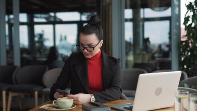 woman in glasses a freelancer, sits in a stylish restaurant with a laptop, dressed in business attire, and glasses, smiling happily upon receiving a message about good news of winning and victory