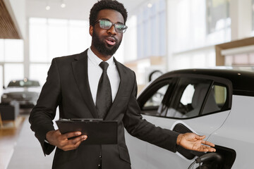 Concept of buying eco-friendly car for progressive customer. Young bearded African salesman, selling electric cars in light modern showroom with panoramic windows.