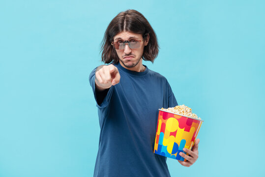 Young long hair tattooed man in casual t-shirt, 3D clip-on glasses (Anaglyph) pointing at camera isolated on blue background studio. Watching movie film hold bucket of popcorn.