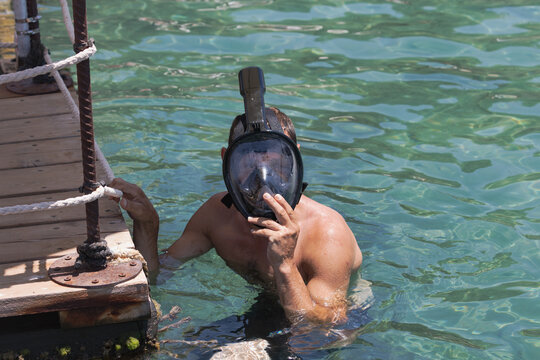 Man in water next to a wooden dock. The person is adjusting a snorkeling mask. Sunlight illuminates the scene creating reflections on the sea surface. Ropes and metal fixtures are visible on the dock
