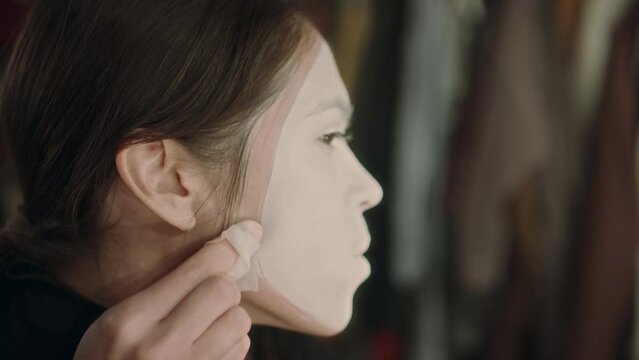 Young female artist using napkin to wipe excesses of white paint on face while creating mime makeup before performance