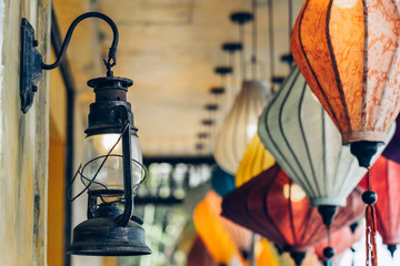 Vintage lantern amidst colorful lanterns. An old-fashioned lantern glows softly, juxtaposed with vibrant, colorful fabric lanterns in the background. - obrazy, fototapety, plakaty