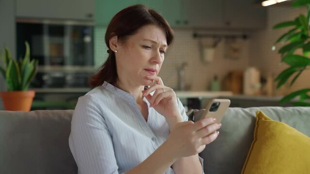 Middle aged woman disappointed looking at smartphone screen. Dissatisfied with bad news message. Unhappy female mobile app user received large bill while sitting on sofa in living room at home