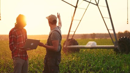 irrigation agriculture. two farmers silhouette with a laptop work in a field with corn at the back...