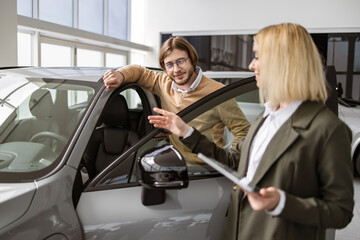 Beautiful woman showing to potential buyer automobile, they looking on car cabin with opened doors. Female car dealer standing with client of dealership in showroom. Manager working with client.