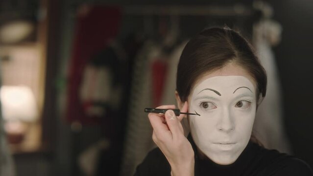 Mime actress drawing black line on white face while applying stage makeup before theatre performance