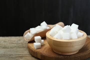 Fototapeta na wymiar White sugar cubes in bowl and scoop on wooden table against dark background, closeup. Space for text