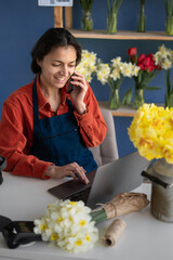 Own business concept. Happy hispanic business woman talking on mobile phone while standing in apron in small floral shop taking order. Joyful female florist calling on smartphone.