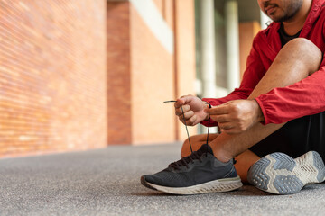 Closeup of sportsman wearing a red jacket tying sneakers outdoors brick wall on city street. Athletic male in hoodie sweatshirt red stopping lacing shoes in urban city park.