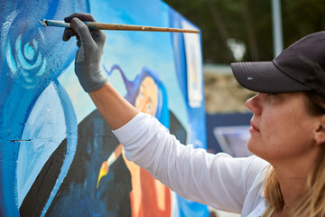 Female painter in black cap draws picture with paintbrush on canvas for outdoor street exhibition, close up side view of female artist apply brushstrokes to canvas, symphony of art creativity