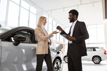 African American sales manager in suit giving car key to young Caucasian blond woman standing...