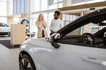Happy husband buying to his loving wife new vehicle at car dealership. African American boyfriend covering eyes of his Caucasian young girlfriend for surprise in car showroom.